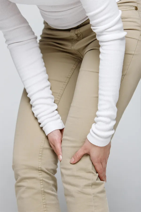 Arthritis-Treatment A woman wearing a white long-sleeve shirt and tan pants, clutching her knee, suffering from arthritis. Get arthritis treatment from Discovery Health Healing Center in Green Bay.