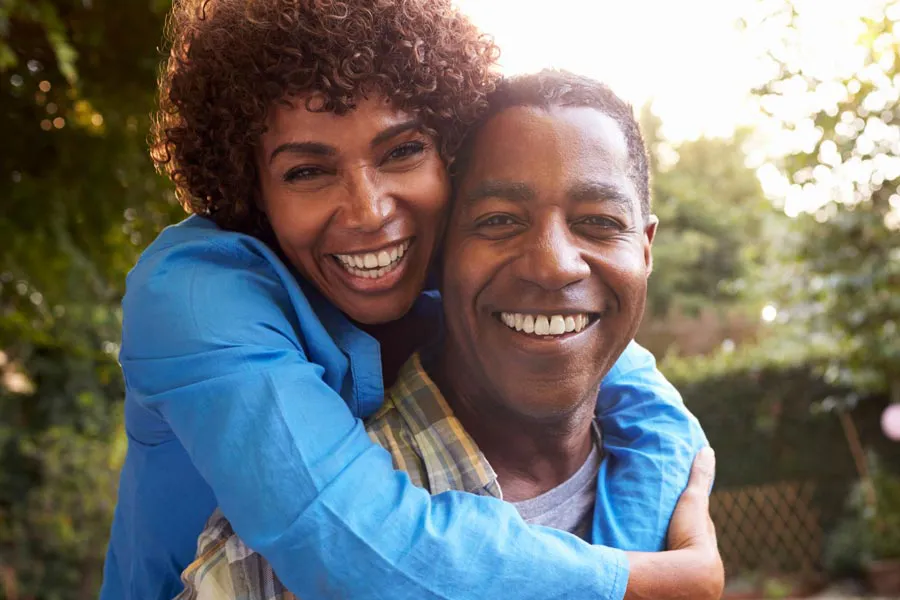 Autoimmune-Doctor A middle-aged black couple outside, the woman embracing the man from behind pressing her cheek to his, both are smiling due to successful Autoimmune Disorder treatment from Discovery Health Healing Center in Green Bay.