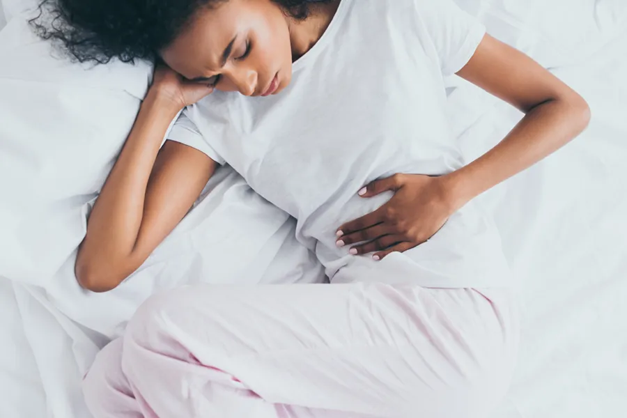 Crohns-Disease-Clinic A dark-skinned woman in white clothes lays on her bed clutching her middle before getting treatment for Crohn's Disease from Discovery Health Healing Center in Green Bay.
