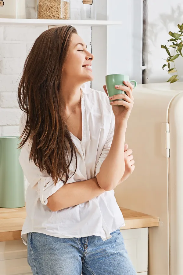 Crohns-Disease-Treatment A woman with a white blouse an jeans drinking tea in her kitchen, smiling after getting treatment for Crohn's Disease from Discovery Health Healing Center in Green Bay.