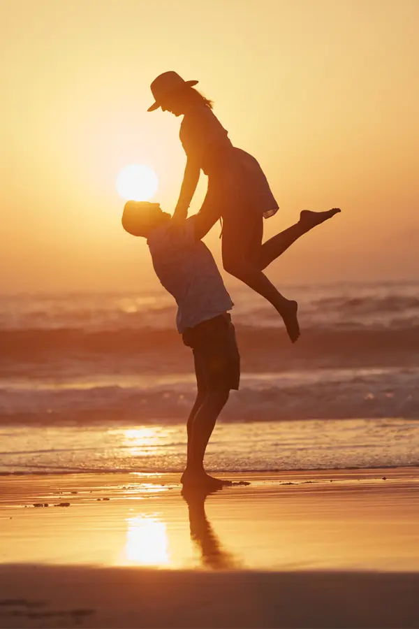 Foot-Detox-Treatment A man lifts his wife into the air on the beach, silhouetted by the sunset. Get Foot Detox treatment from Discovery Health Healing Center in Green Bay.