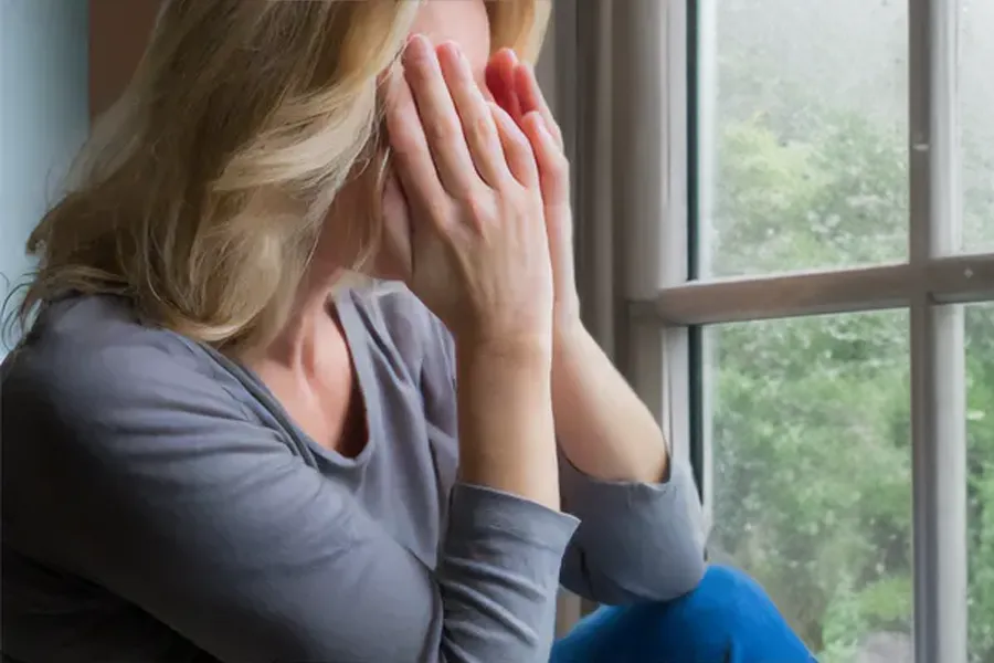 Graves-Disease-Clinic A blonde woman in a gray shirt sits in front of a window on a rainy day covering her eyes with her hands. Get treatment for Grave's Disease from Discovery Health Healing Center in Green Bay.
