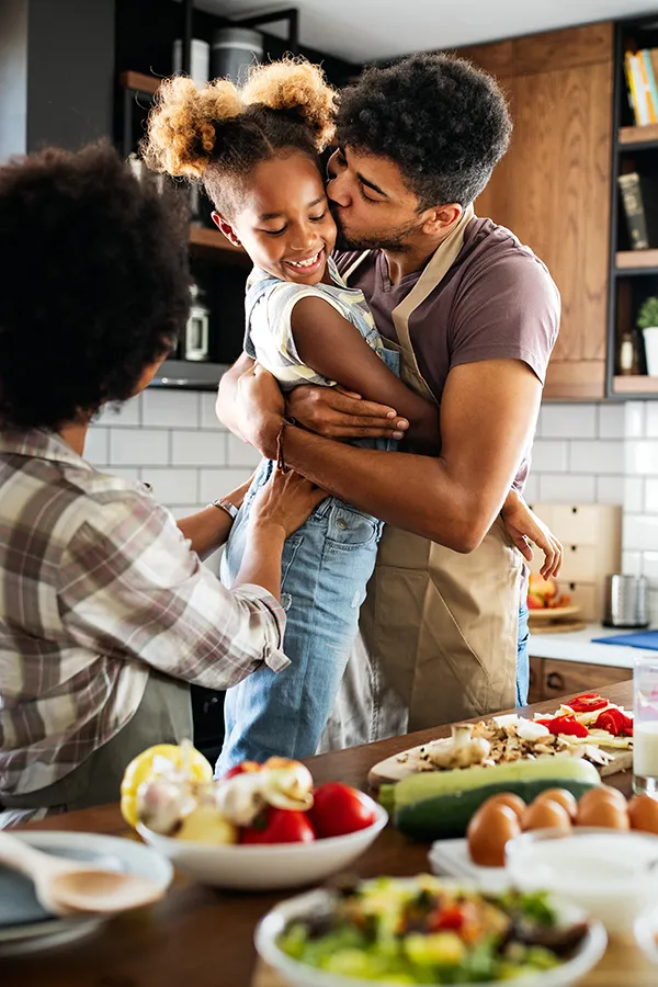 Gut-Health-Treatment A man holds his daughter and kisses her cheek as the family makes a healthy dinner in the kitchen. Get help for digestive issues and gut health from Discovery Health Healing Center in Green Bay.