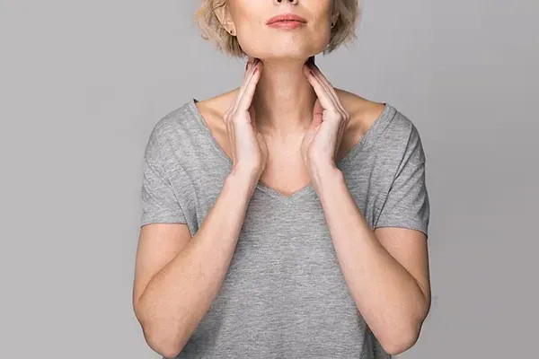Hashimotos-Thyroiditis-Doctor An older blonde woman in a gray t-shirt holds her hands to her neck checking for signs of thyroid problems. Get help for Hashimoto's Thyroiditis from Discovery Health Healing Center in Green Bay.