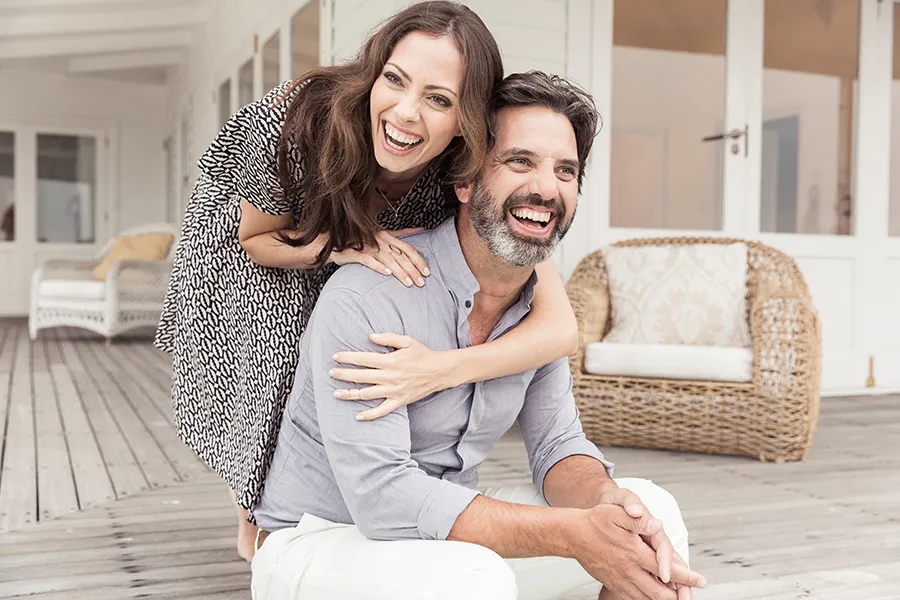 A middle-aged couple on a wooden porch with wicker furniture; the woman is playfully hugging the man from behind. They are benefiting from the health and wellness services from Discovery Health Healing Center in Green Bay.