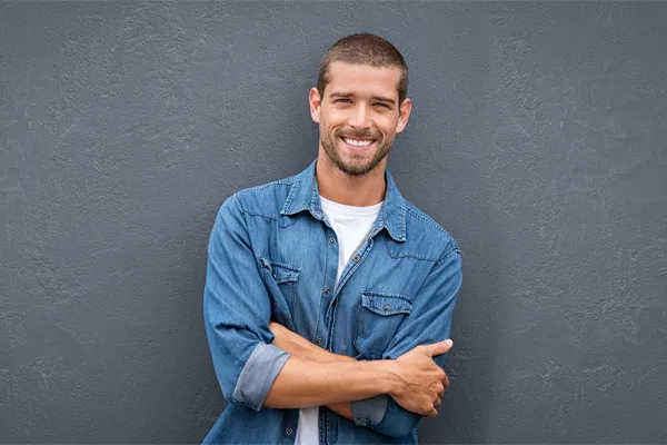 Hormones-for-Men-Doctor A man in a denim shirt stands smiling against a gray-blue wall, pleased with his testosterone hormone therapy from Discovery Health Healing Center in Green Bay.