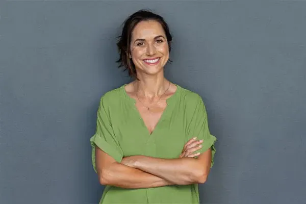 Hormones-for-Women-Doctor A woman in a green shirt stands smiling against a gray-blue wall, pleased with her hormone therapy from Discovery Health Healing Center in Green Bay.