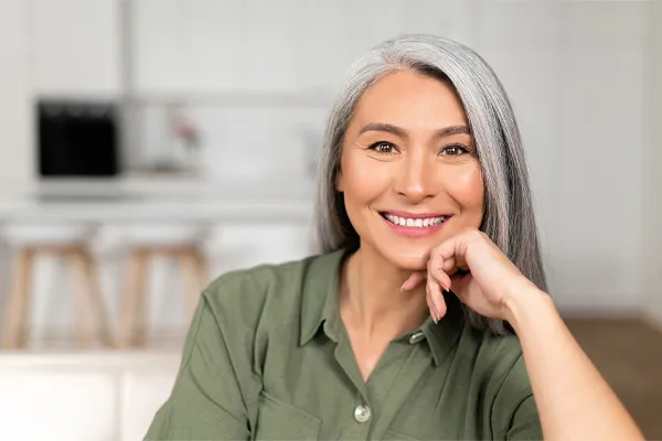 Menopause-Clinic An asian woman with long gray hair and a green shirt sitting and smiling on her couch, happy with her menopause care from [NAME} in Green Bay.