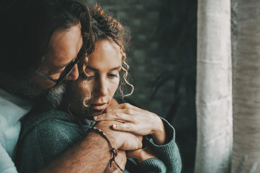 Neurodegenerative-Disorders-Therapy A man embraces a woman from behind in a dimly lit room, their somber expressions showing concerns about a neurodegenerative disease they will get treated by Discovery Health Healing Center in Green Bay.