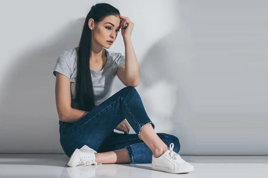 PCOS-Doctor A young brunette woman in a gray t-shirt and jeans sits against a gray wall with her head resting on her hand and a distressed expression. Get help for Polycystic Ovarian Syndrome (PCOS) from Discovery Health Healing Center in Green Bay.