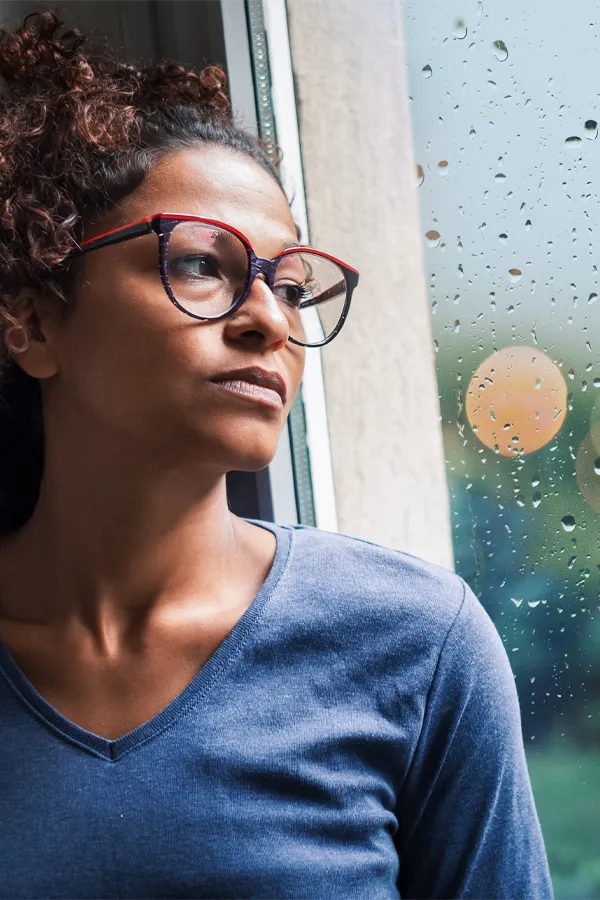 PMDD-Treatment A black woman with with a somber expression and glasses and a gray shirt stares out the window on a rainy day. Get treatment for Premenstrual Dysphoric Disorder from Discovery Health Healing Center in Green Bay.
