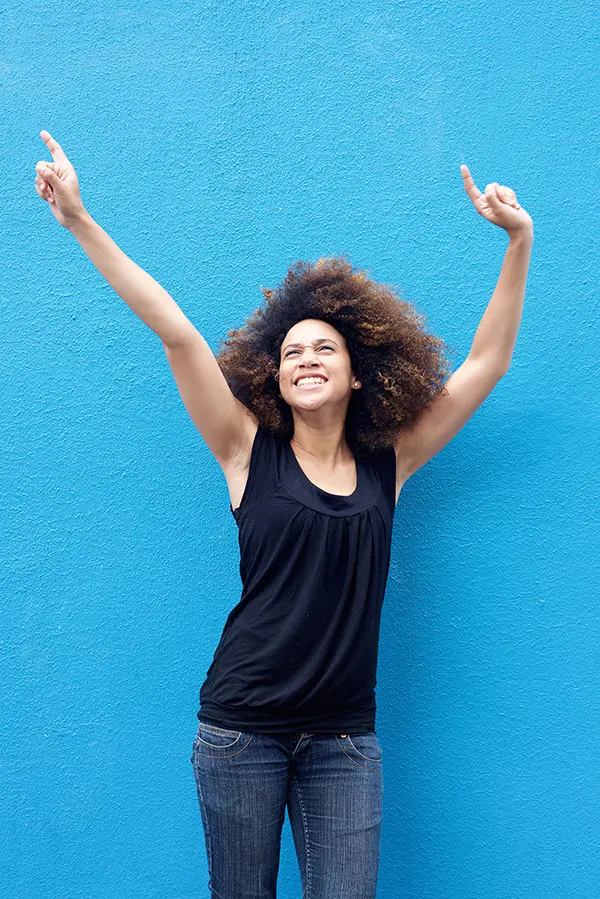 PMS-Treatment A woman in a dark blue tank top standing in front of a bright blue wall, raising her arms in celebration of relief from PMS from Discovery Health Healing Center in Green Bay.